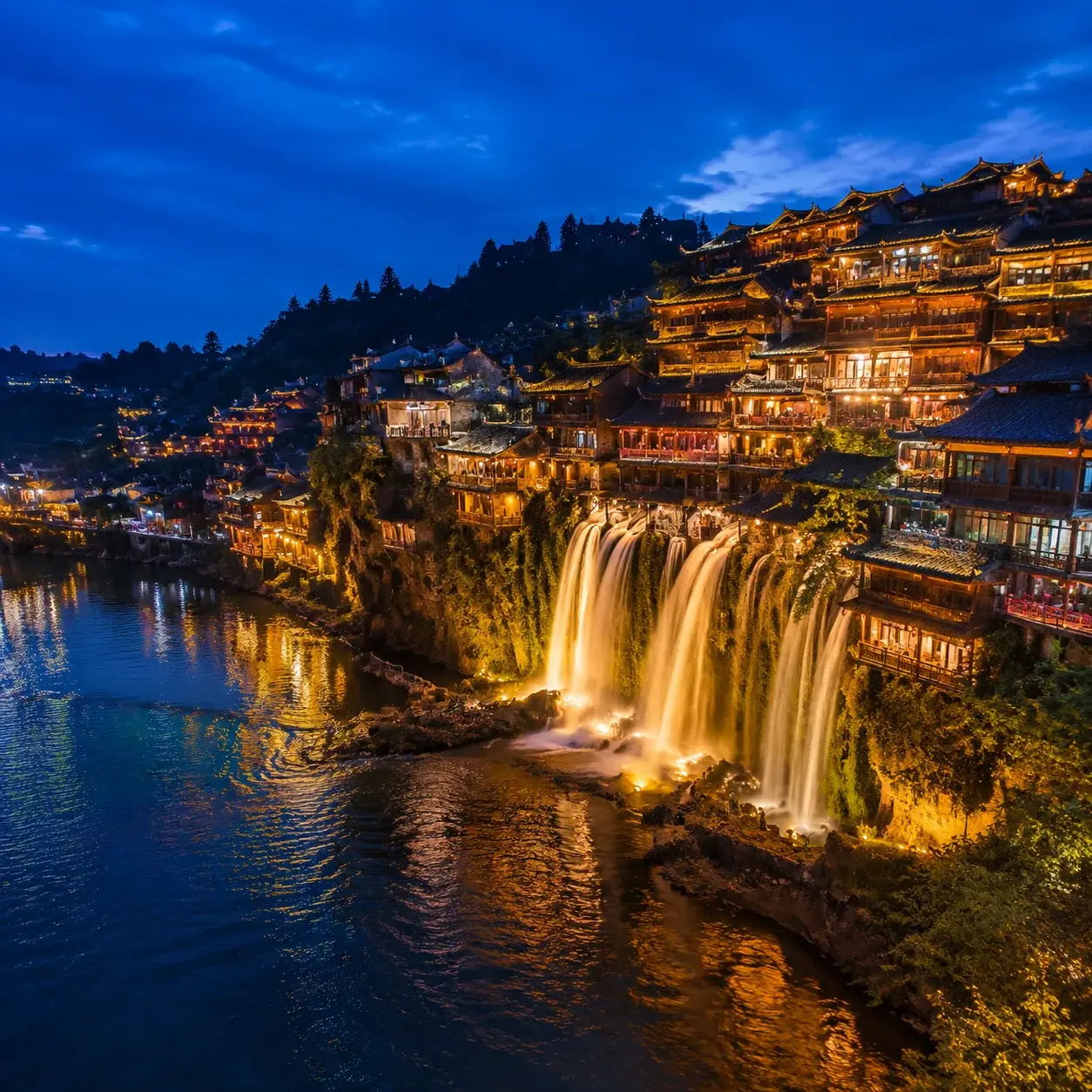 Furong Ancient Town glowing with warm lights above its illuminated waterfall at blue hour