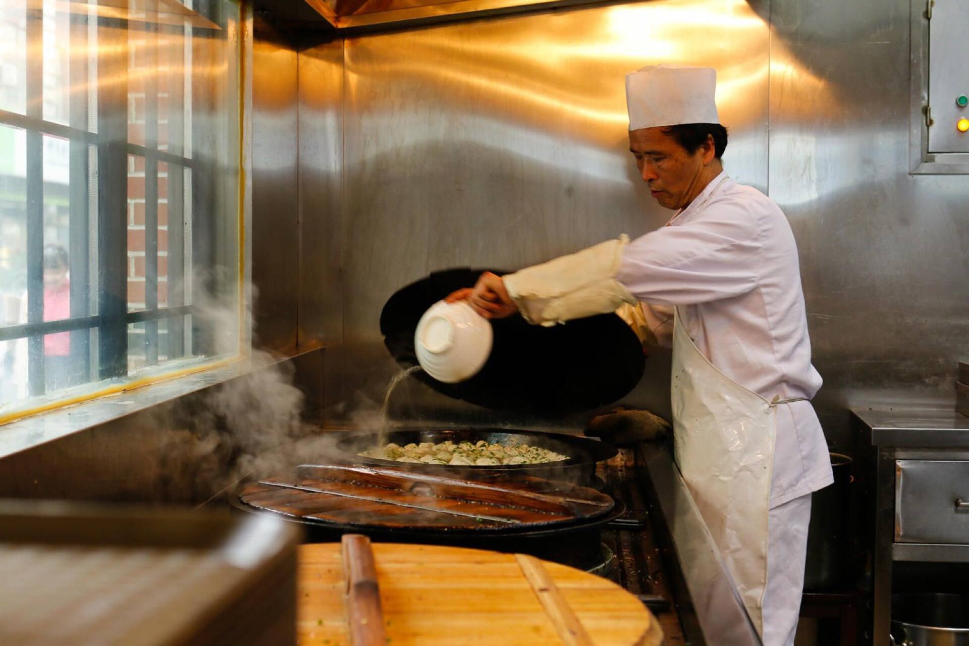 A cook pouring broth into a pan of frying buns
