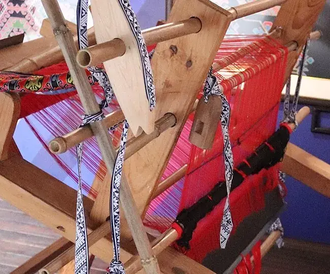 Red warp under tension on the slant loom, the weaver's back-strap hanging beside it