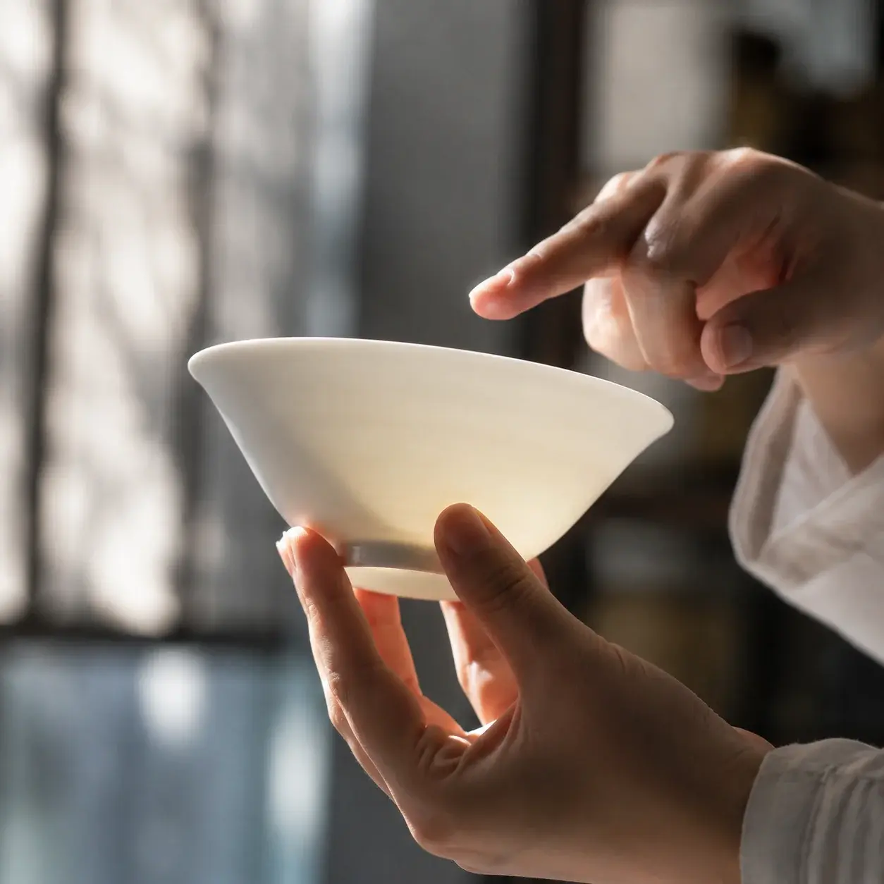 Hands hold a thin porcelain bowl to the light, showing its faint translucency.