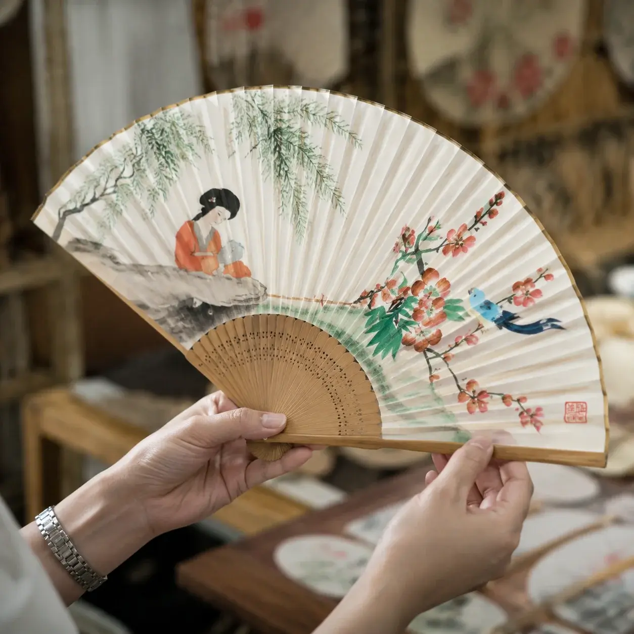 Hands hold an open silk folding fan close up, revealing brush-painted details, a red seal, and fine bamboo ribs.