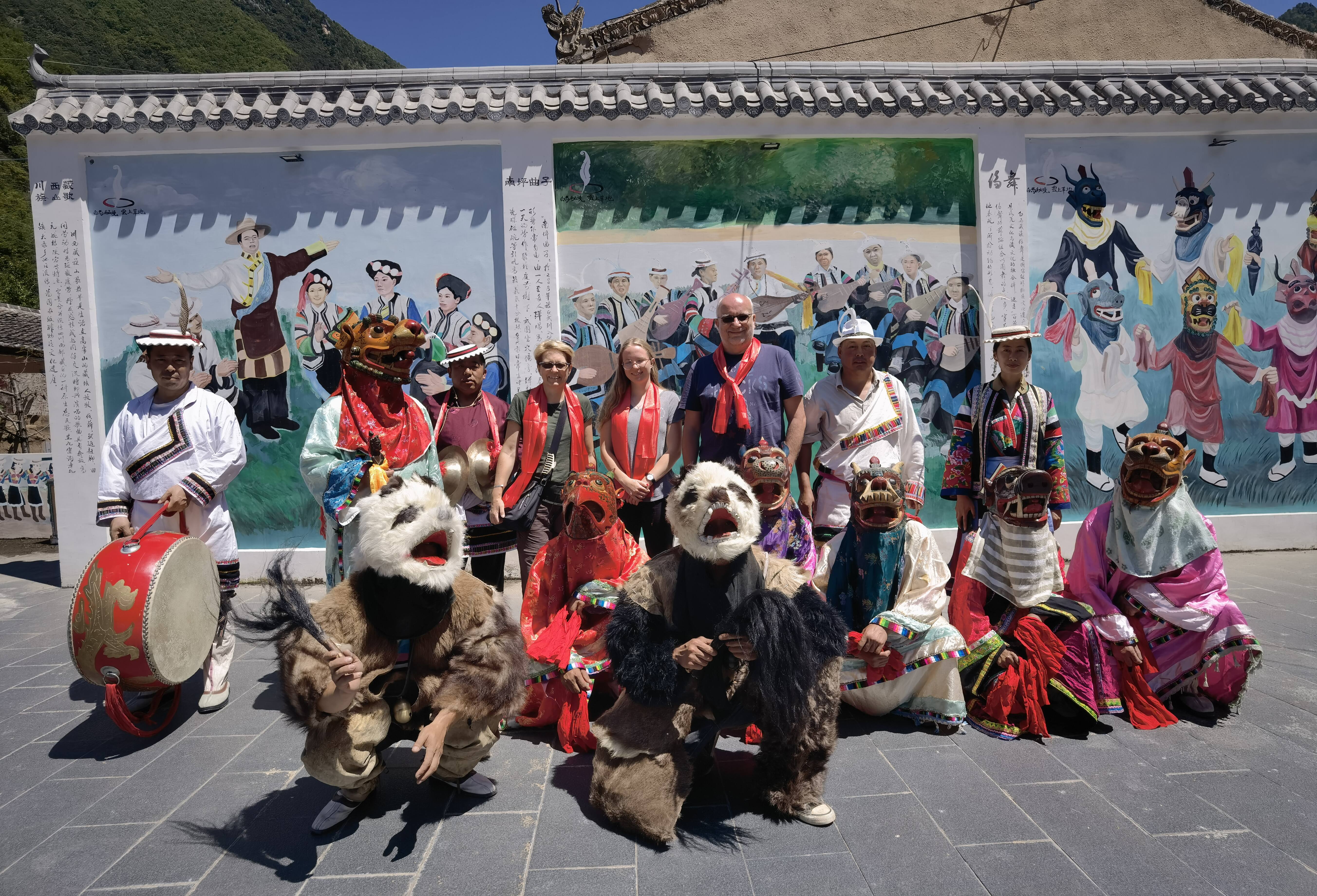 Guests with red scarves standing with panda-costume dancers and masked performers in front of a village mural